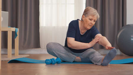 Senior woman stretching sitting on yoga mat in living room. Active healthy lifestyle sporty old person training workout home wellness and indoor exercisingの写真素材