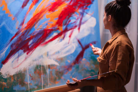 Professional woman painter looking at painting and holding paint palette. Modern artwork paint on canvas, creative, contemporary and successful fine art artist drawing masterpieceの写真素材