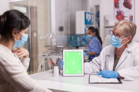 Doctor with face mask looking at tablet with green screen during consultation with sick patient. Nurse wearing blue uniform.の写真素材