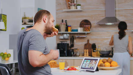 Entrepreneur on a video conference while eating breakfast in kitchen. Freelancer working remote, talking in videoconference video call online web internet meeting from home, communication deviceの写真素材