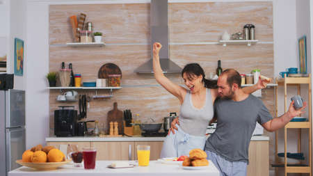 Carefree happy and joyful couple dancing and singing in kitchen in synny morning. Cheerful husband and wife laughing, singing, dancing listening musing, living happy and worry free. Positive peopleの写真素材
