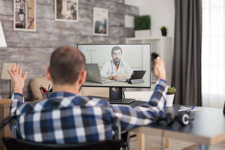 Patient on video call with doctor talking about his rehabilitation. Young immobilized guy sitting at home in front of laptop, talking with his doctor about the process of rehabilitation on video call.の写真素材