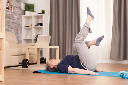 Old woman exercising in her apartment sitting on yoga mat. Old person pensioner online internet exercise training at home sport activity with dumbbell, resistance band, swiss ball at elderly retirement ageの写真素材