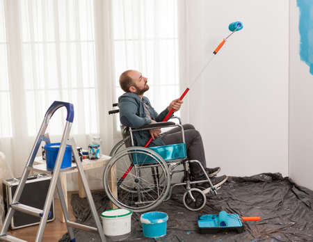Invalid man painting wall while sitting in wheelchair. Handicapped, disabled ill and immobilizes man...