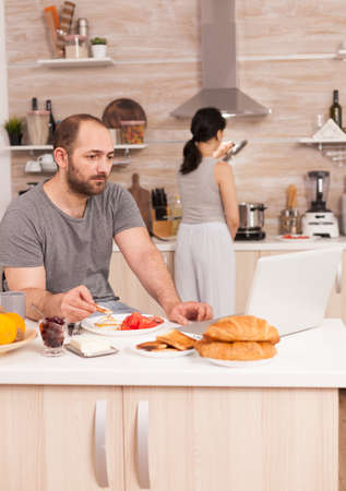 Entrepreneur working from home while eating breakfast in kitchen, wearing pajamas enjoying roasted bread with butter. Freelancer working online via internet using modern digital technologyの写真素材