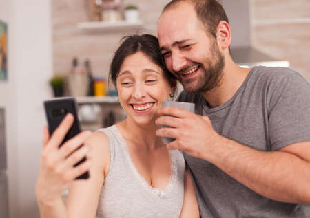Amused couple looking at the phone in the morning during the breakfast. Joyful married husband and wife making funny faces while taking a photo during breakfast in kitchen.の写真素材