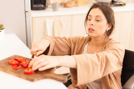 Woman with walking handicap cutting red paper on wooden board in kitchen using sharp knife.の写真素材