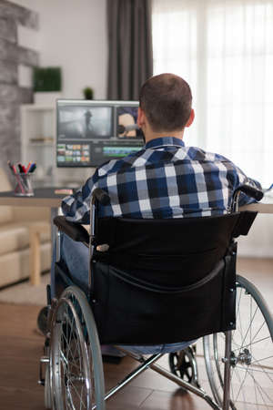 Disabled video editor sitting on wheelchair doing post production on film. Invalid videographer doing his business online, using high technology, sitting in his apartment, working remotely in special conditions.の写真素材