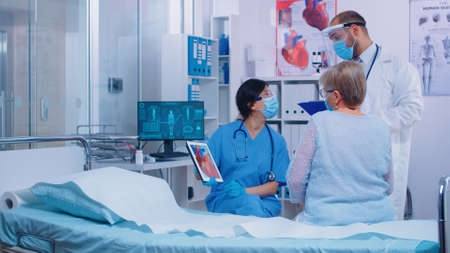 Nurse in mask with cardiac patient at doctor appointment showing a heart representation on digital tablet in modern clinic. Doctor entering hospital room talking with old retired senior woman patientの写真素材