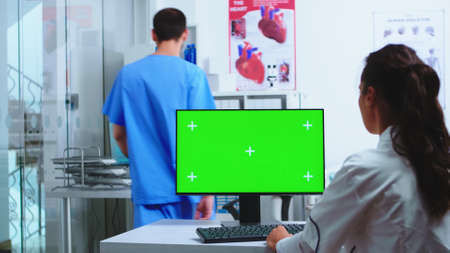 Physician using computer with chroma key and assistant holding patient x-ray in hospital cabinet. Desktop with replaceable green screen in medical clinic while doctor is checking patient radiography for diagnose.の写真素材