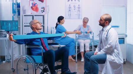 Senior man in wheelchair exercising muscle strength with elastic band under medical supervision in...