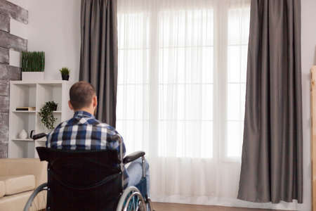 Paralyzed young man sitting alone on wheelchair in living room. Serious sad caucasian man wearing casual clothes and look at large panoramic view in bright modern living room, hospital or clinic.の写真素材