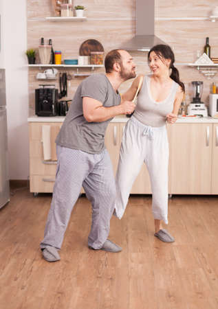 Joyful couple dancing and singing during breakfast in kitchen wearing pajamas. Carefree wife and husband laughing having fun funny enjoying life authentic married people positive happy relationの写真素材