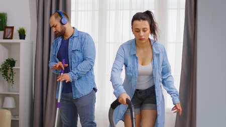 Happy man listening music on headphones and cleaning the floor with mop while his wife is using...
