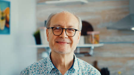 Close up of smiling senior man in kitchen looking at camera wearing eyeglasses. Portrait of relaxed...
