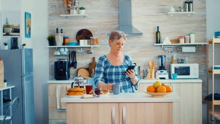 Senior woman browsing on mobile in kitchen during breakfast and smiling to the camera. Authentic...