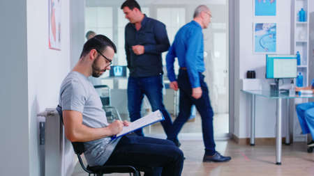 Young man filling documents in hospital waiting area. Nurse inviting senior woman inside examination...