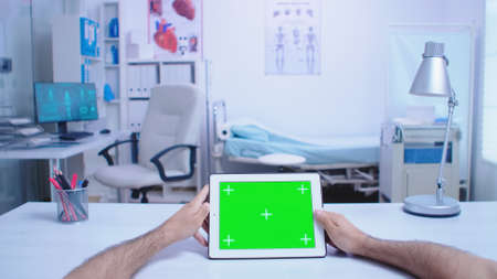 Pov shot of assistant holding tablet pc with green screen in hospital cabinet and doctor wearing white coat working on computer. Medic using tablet with chroma key on display in medical clinic.の写真素材