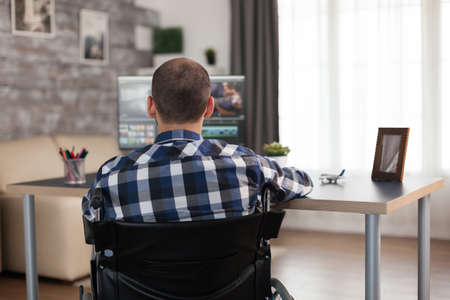 Video editor in wheelchair working on film post production. Invalid videographer doing his business online, using high technology, sitting in his apartment, working remotely in special conditions.の写真素材