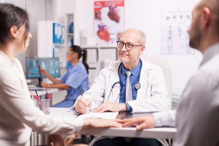 Senior doctor writing notes on clipboard during examination of young couple with fertility illness in hospital office and nurse holding patient x-ray.の写真素材