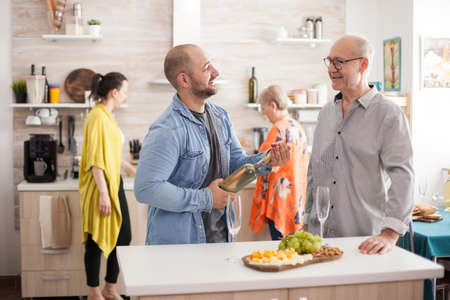 Young man holding a wine bottle while talking with his old dad in kitchen. Mother preparing lunch for family.の写真素材