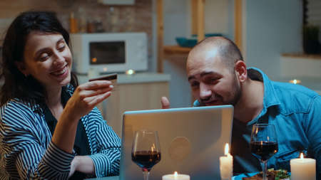 Husband and wife shopping online and making payments during dinner using card. Couple sitting at the table, searching, browsing, surfing, using technology, enjoying festive meal in the dining roomの写真素材