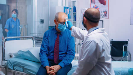 Doctor checking old man temperature with a infrared thermometer. Retired senior man wearing a mask and healthcare worker in protective equipment for consultation. Modern private clinicの写真素材
