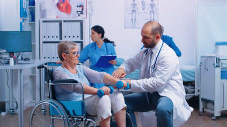 Doctor helping disabled senior patient to recover muscle strength in private modern rehabilitation clinic or hospital. Nurse in background talking with elderly man with walking frameの写真素材