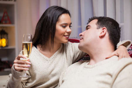 Man looking into her wife eyes sitting on couch celebrating christmas. Woman holding a glass of...