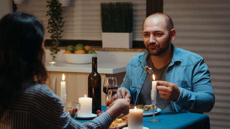 Couple eating and drinking wine with man in foreground during festive dinner in the kitchen. Talking happy sitting at table dining room, enjoying the meal at home having romantic time at candle lightsの写真素材