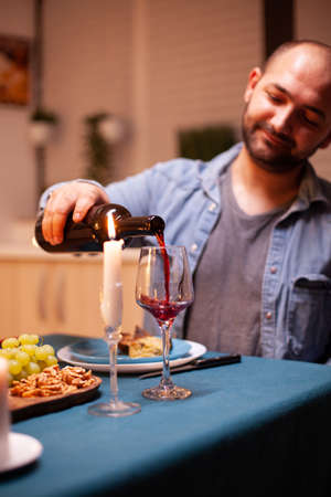 Husband pouring red wine in glass celebrating relationship with wife in dining room. Young man...