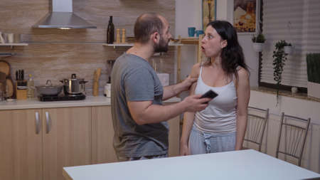 Cheated man yelling at wife and hitting her. Jealous husband angry, frustrated, offended and irritated accusing woman of infidelity arguing her with messages on smartphone screaming desperateの写真素材