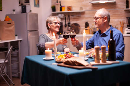 Candles burning on kitchen table and old couple toasting with glasses for red wine for their relationship. Happy cheerful senior elderly couple dining together in the cozy kitchen, enjoying the meal.の写真素材