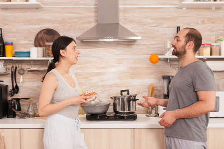 Husband playing with an orange and talking with his wife in the kitchen. Husband having a conversation with wife while she is preparing eggs for breakfast.の写真素材