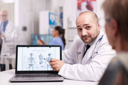 Doctor telling senior patient she needs back surgery during radiography examination in hospital office. Nurse in blue uniform holding x-ray in the background. Elderly physician in clinic corridor.の写真素材