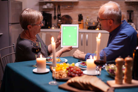 Green screen tablet at dinner. Aged people looking at green screen template chroma key display sitting at the table in kitchen during dinner.の写真素材