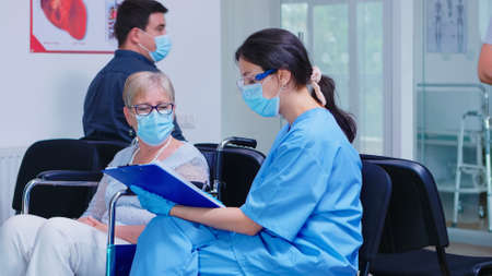 Nurse explaining diagnosis to disabled senior woman in wheelchair. Assistant wearing face mask against infection with coronavirus in hospital waiting area.の写真素材