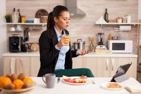 Businesswoman having an online meeting in the morning. Freelancer reading the last news online before going to work, using modern technology in the kitchen while eating a healthy meal.の写真素材