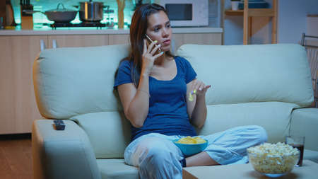 Speaking on phone holding snacks bowl and eating snacks in front of TV. Happy, relaxed, lonely lady in pajamas enjoying the evening sitting on comfortable couch in front of television using smartphoneの写真素材