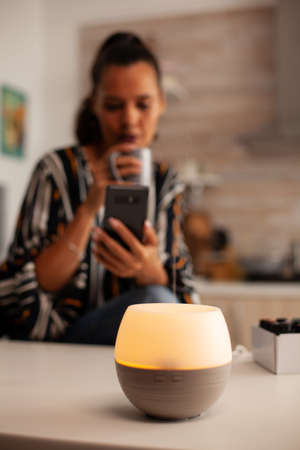 Woman browsing on phone enjoying aromatherapy from essential oil diffuser. Aroma health essence, welness aromatherapy home spa fragrance tranquil theraphy, therapeutic steam, mental health treatmentの写真素材