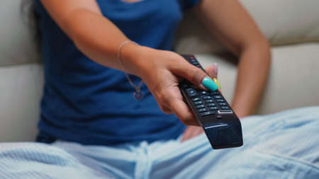Close up of switching tv programs sitting on sofa using remote control. Shot of controller in hands of person pointing to TV pressing the button and changing channels sitting in front of televisionの写真素材