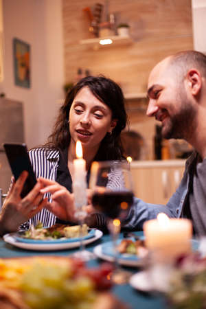 Wife showing husband photos on smartphone during festive relationship anniversary. Adults sitting at...