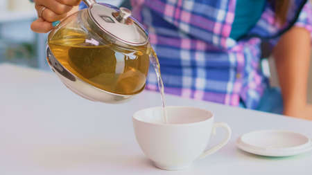 Close up of tea from the kettle slowly pour into porcelain cup. Young lady preparing green tea in the kitchen in the morning at breakfast, using teapot, teacup and healty herbal leaves.の写真素材