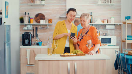 Woman showing a funny movie on smart phone to her mother, sitting in the modern kitchen around the table drinking a glass of white wine. Senior person learning browsing using internet technologyの写真素材