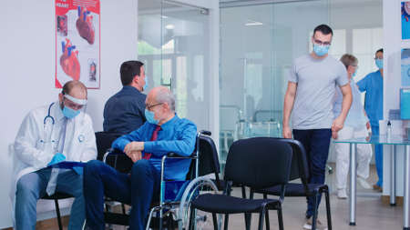 Invalid old man with face mask against infection with coronavirus in wheelchair discussing with doctor in hospital waiting area. Nursing inviting patient in examination room.の写真素材