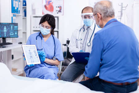 Doctor and nurse explaining radiography to senior man during consultation on tablet pc in hospital with protection mas against covid-19. Medical examination for infections, disease and diagnosis.の写真素材