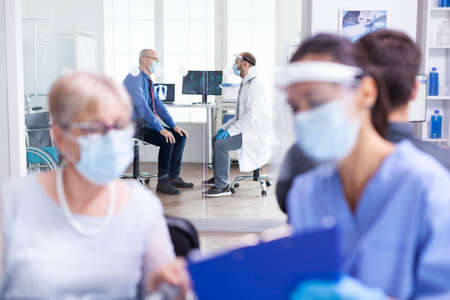 Elderly man discussing with doctor in hopspital examination room wearing protection against...