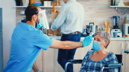 Social worker with face mask taking body temperature of disabled senior woman in wheelchair during home visit and coronavirus pandemic. Geriatrician helping prevent covid-19 spreadの写真素材