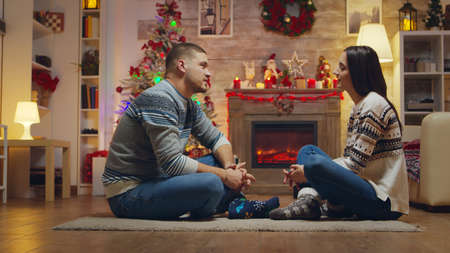 Beautiful young couple looking at each other sitting on the carpet celebrating christmas in front of fireplace.の写真素材