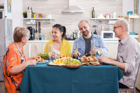 Mother and daughter smiling while having delicious lunch in kitchen with family. Senior man holding wine glass. Tasty roasted potatoes.の写真素材